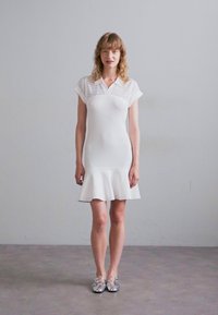 Abito bianco aderente con maniche corte, dettagli a occhio di pernice sulle spalle, texture a costine e orlo svasato. Il modello indossa scarpe argentate.