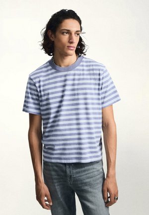 Ung mand med skulderlangt mørkt hår iført en lyseblå og hvidstribet t-shirt og falmede blå jeans, der kigger til siden.