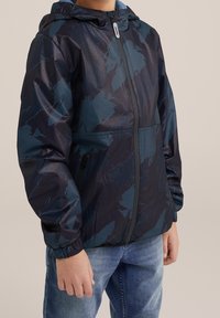 Dětská bunda v černé a modré barvě s texturovaným povrchem, zipovým uzávěrem, kapucí a elastickými manžetami; má jemný geometrický vzor.