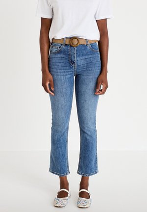 Person iført lyseblå, korte flare-jeans med et vævet bælte, hvid T-shirt og blomstrede Mary Jane-sko, der står mod en ensfarvet baggrund.