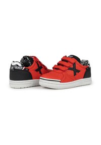 Zapatillas rojas con acentos negros, cuello acolchado y correas de Velcro; textura perforada en la parte superior, suela blanca y detalle de logo distintivo.