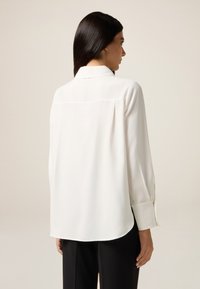 Weiße Langarmbluse aus glattem Stoff, mit klassischem Kragen, Knopfmanschetten und abgerundetem Saum. Minimalistisches Design, ohne Muster.
