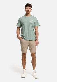 T-shirt en coton vert clair avec un graphique à l'avant, associé à un short beige à rayures et des baskets montantes blanches. Le modèle se tient dans une pose neutre.