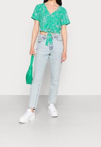 Top verde a maniche corte con stampa di foglie e nodo frontale, abbinato a jeans a vita alta blu chiaro e sneakers platform bianche.