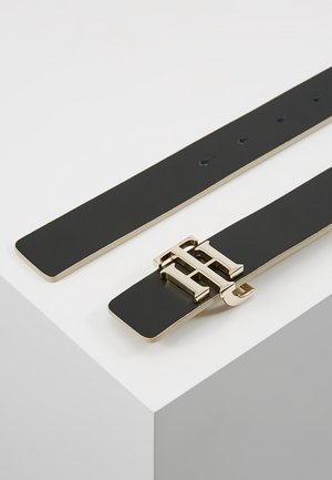 Czarny skórzany pasek z podwójną złotą klamrą w kształcie litery "T", ułożony płasko na białej powierzchni, z widocznymi wieloma otworami do regulacji.