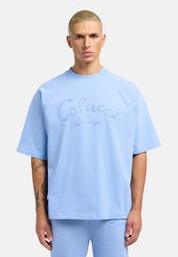Light blue short-sleeve sweatshirt with a crew neck, featuring embroidered text "Colucci la Famiglia" in varying shades of blue.