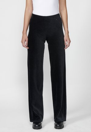 Mujer con pantalones negros de cintura alta y perneras anchas con una textura sutil, combinados con zapatos negros y una camiseta blanca sin mangas.