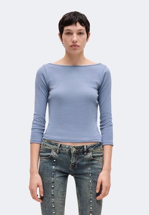 Jeune personne aux cheveux courts et foncés portant un haut bleu clair à manches longues et un jean foncé avec des détails de perles verticales.