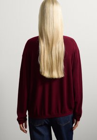 Personne avec de longs cheveux blonds raides portant un pull bordeaux et un jean bleu, se tenant face à un mur gris clair uni.