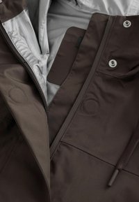 Veste marron foncé avec fermeture éclair partiellement ouverte, boutons-pression argentés, doublure intérieure grise et cordon marron foncé visible.