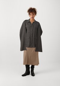 Jachetă oversized gri cu guler și patru buzunare frontale, asortată cu o fustă de satin bej și cizme negre până la genunchi.
