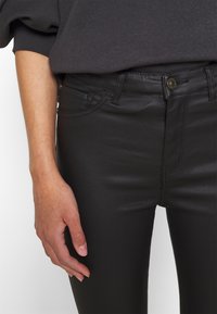 Svarta högmidjade jeans med en slät yta, detaljerad sömnad och en framknäppning. Handen vilar på midjebandet.