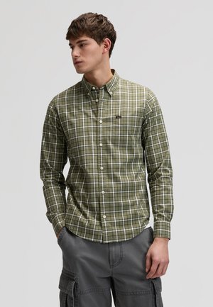 Młody mężczyzna w zielono-białej koszuli w kratę na guziki i szarych spodniach typu cargo, patrzący w lewo, z rękami w kieszeniach.