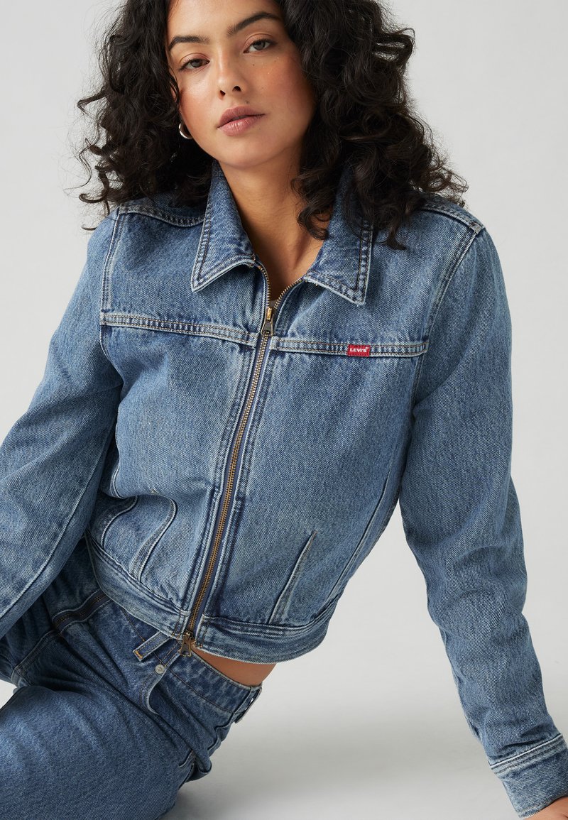 Chaqueta de mezclilla en azul claro con cremallera frontal, cuello puntiagudo y dos bolsillos en el pecho. Presenta un pequeño parche rojo con el logo de Levi's en el lado izquierdo.