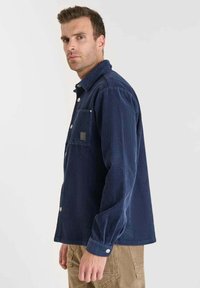 Camicia in corduroy blu marina con maniche lunghe, chiusura a bottoni, taschino sul petto e dettagli in metallo argentato. Tessuto testurizzato con linee verticali.