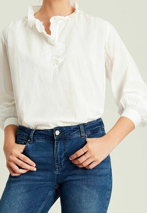 Kobieta ubrana w białą bluzkę z marszczonym kołnierzem i długimi rękawami oraz dopasowane niebieskie dżinsy, ręce opierające się na biodrach na jasnym tle.