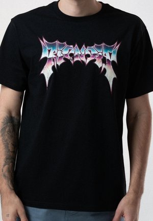 Mężczyzna w czarnej koszulce z kolorowym, stylizowanym napisem „Veneno” na klatce piersiowej oraz widocznym tatuażem lwa na lewym przedramieniu.