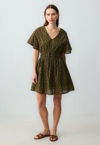 Olivengrünes Spitzenkleid mit V-Ausschnitt, kurzen Flatterärmeln, elastischem Bund und floraler Lochmusterung; getragen mit braunen Riemchensandalen.