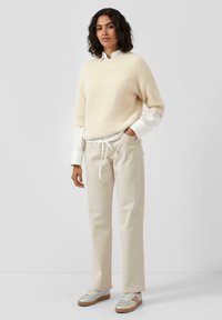 Beżowy sweter z dzianiny, pod spodem biały koszula z kołnierzykiem, kremowe spodnie z troczkiem i srebrne sneakersy z beżową podeszwą. Kręcone włosy okalające twarz.