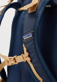 Jūrų mėlynumo audinio kuprinė su smėlio spalvos diržais, kuriame yra juodas Patagonia logotipas ir plastikinių sagtelių aparatas. Lygi tekstūra ir paminkštinta konstrukcija.