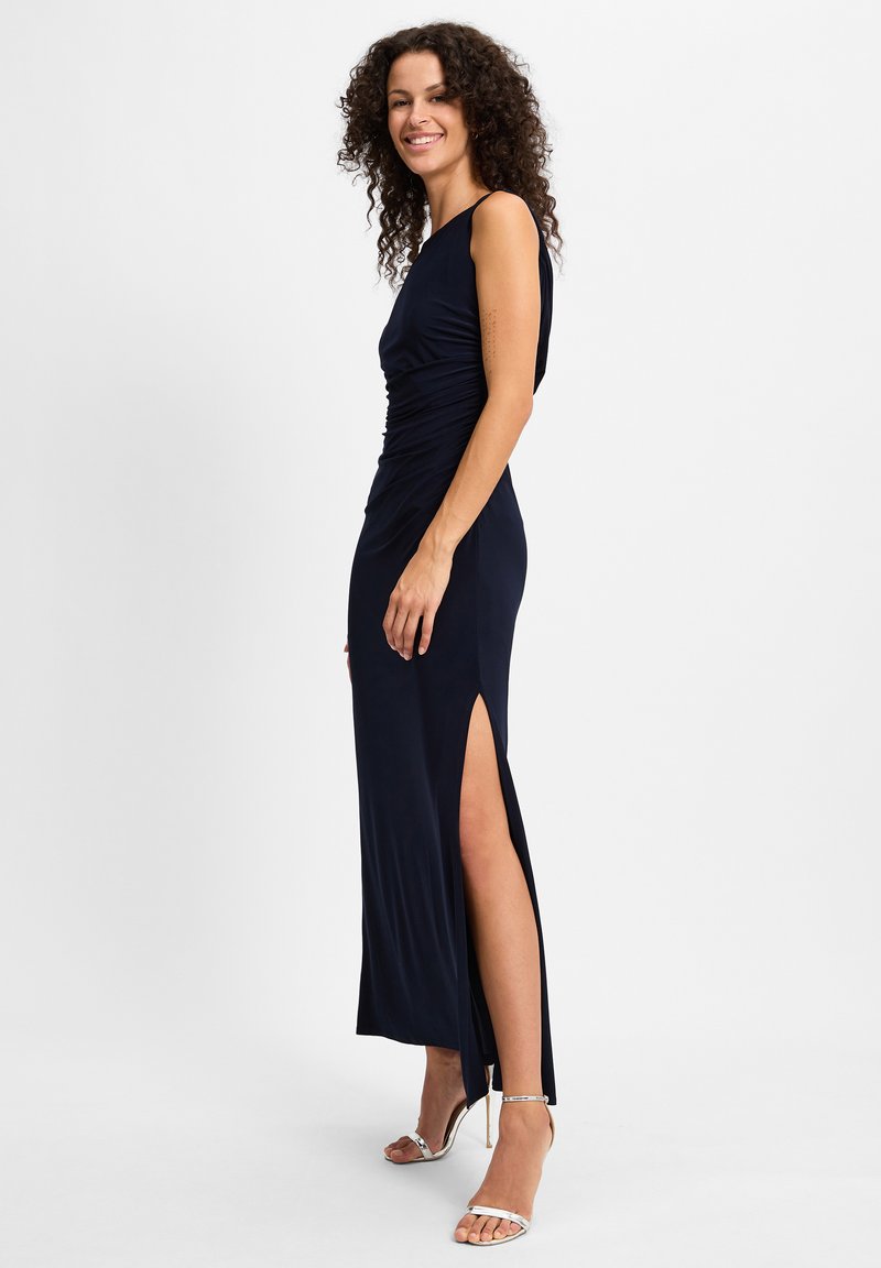 Femme dans une robe sans manches bleu marine avec une fente latérale haute, portant des sandales à talons hauts argentées, debout et souriant sur le côté.