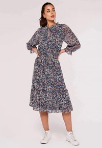 Blumenkleid in Marineblau, mit Rüschensaum, langen Ärmeln, elastischem Bund und mehrlagigem Saum. Kombiniert mit weißen Sneakers.