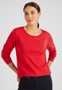 Rode pullover trui van zacht materiaal, met een ronde halslijn en lange mouwen. Het heeft een decoratief patroon van rhinestones aan de ene kant.