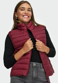 Burgundy pufferväst med hög krage och vertikal quiltad design, ihop med en svart tröja och grå högmidjade jeans.