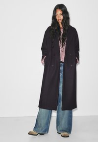 Donna con capelli lunghi e scuri, indossante un lungo cappotto nero, una maglietta a righe rosse e bianche, jeans a gamba larga e stivali marrone chiaro, in piedi contro un muro bianco.