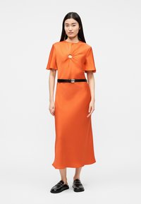 Γυναίκα που στέκεται, φοράει μία φωτεινή πορτοκαλί μίντι φόρεμα με κοντά μανίκια, κεντρικό άνοιγμα στο στήθος, μαύρη ζώνη και μαύρα παπούτσια χωρίς δέσιμο.