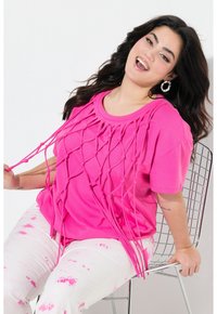 Camiseta rosa de gran tamaño con un diseño de trenzado anudado en la parte frontal, mangas cortas, combinada con pantalones blancos que presentan patrones de salpicaduras rosas.