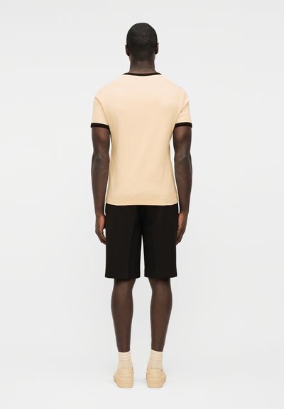 Άνδρας στέκεται με την πλάτη προς τα εμπρός, φοράει μπεζ μπλούζα με κοντά μανίκια και μαύρο περίγραμμα, μαύρο σορτς, μπεζ κάλτσες και μπεζ αθλητικά παπούτσια, μπροστά σε λευκό φόντο.