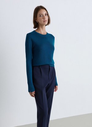 Donna con capelli corti castani che indossa un maglione verde acqua a maniche lunghe e pantaloni blu scuro, in piedi davanti a uno sfondo chiaro e uniforme.