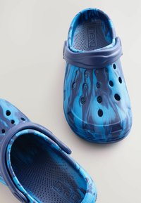 Blå mönstrade träskor med en texturerad yta, som har ett rem, ventilationshål och en slät insida. Tillverkade av flexibelt plastmaterial.