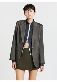 Blazer in tweed grigio con spalle strutturate, bottone oro singolo, indossato sopra un top a zip rigato blu scuro e una gonna verde oliva.