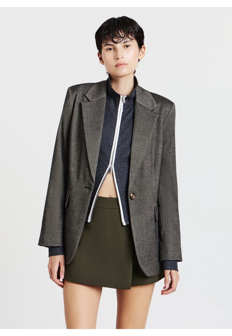 Blazer in tweed grigio con spalle strutturate, bottone oro singolo, indossato sopra un top a zip rigato blu scuro e una gonna verde oliva.
