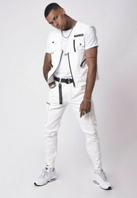 Pantalones cargo blancos con bolsillos con cremallera, un chaleco sin mangas a juego y una camiseta blanca debajo. Las zapatillas también son blancas con detalles en negro.
