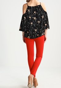 Haut noir à épaules nues avec motif floral, manches évasées et encolure élastique, associé à un jean rouge ajusté et des talons beiges.