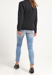 Sudadera negra con puños acanalados, combinada con jeans ajustados de color azul claro y zapatos negros tipo slip-on, vista desde la parte de atrás.