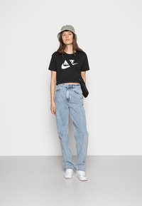 Camiseta negra corta con logo blanco de Nike, vaqueros holgados de color azul claro, sombrero de cubo gris y zapatillas blancas con detalles en rosa. Bolso cruzado negro.