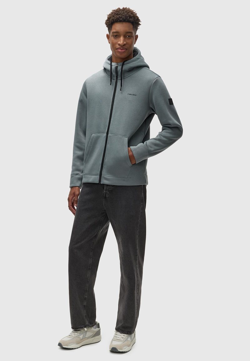 Grauer Zip-Hoodie mit Kapuze, Fronttaschen und schwarzen Akzenten, kombiniert mit dunklen Jeans und grauen Turnschuhen. Glatte Textur.