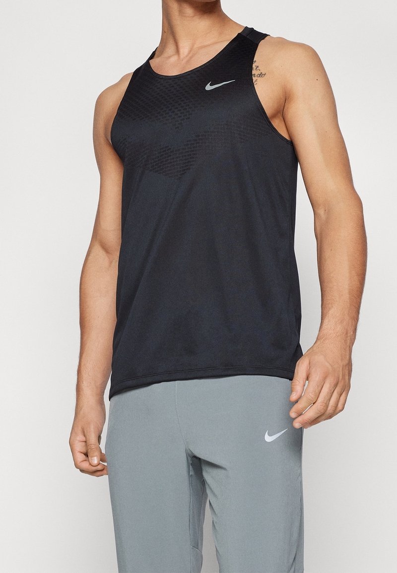 Homme portant un débardeur sportif noir et un pantalon de sport ajusté gris clair avec des logos Nike sur les deux vêtements.