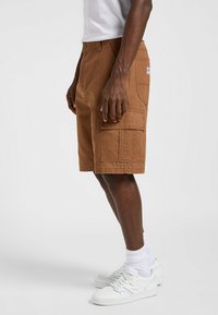 Shorts cargo marrones hechos de tela duradera, con múltiples bolsillos, un ajuste holgado y detalles de costura visibles. Llevados con zapatillas blancas.