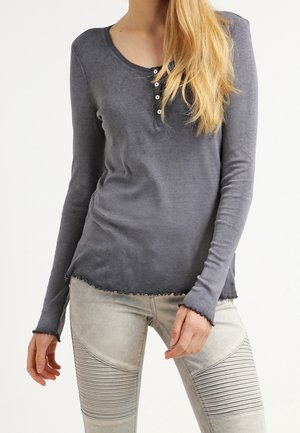 Femme portant un t-shirt henley ajusté à manches longues gris foncé avec des boutons et un pantalon texturé gris clair, debout, les mains près des hanches.