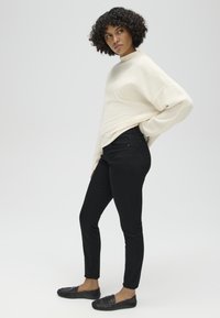 Vrouw met krullend haar draagt een crèmekleurige trui, zwarte broek en zwarte loafers, staand met één hand op haar heup tegen een effen achtergrond.