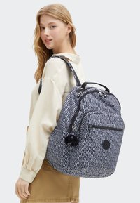 Marinblå och vit mönstrad ryggsäck med flera fack, dragkedjor och en liten svart pom-pom charm. Tygliknande material, rektangulär form.