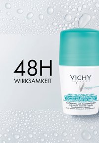 Vichy anti-transpirant, white cylindrical container with a turquoise cap. Text mentions 48-hour effectiveness. Surrounded by water droplets.