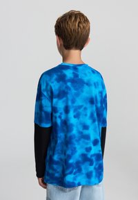 T-shirt a maniche corte con fantasia tie-dye blu a effetto sfumato. Il tessuto sembra morbido, abbinato a un livello a maniche lunghe nero sotto.