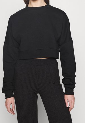 Femme portant un pull court noir à manches longues et un pantalon en maille côtelée noir taille haute sur un fond clair uni.