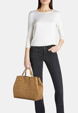 Donna con top bianco a maniche lunghe e jeans neri che tiene una grande borsa in pelle beige con decorazioni in oro.
